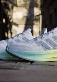 Chaussures de course blanches avec semelles rembourrées bleu clair et jaune sur une rue pavée dans un environnement urbain.