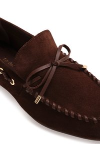 Mocassin en daim marron foncé avec lacet en cuir noué en noeud et embouts métalliques, présentant des détails en cuir cousus le long du bord.