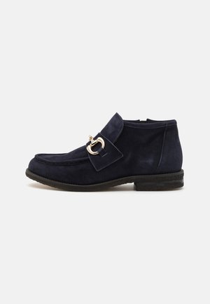 Marineblaue Wildleder-Stiefeletten mit runder Zehenpartie, ausgestattet mit einer dekorativen goldenen Schnalle und einer flachen Sohle, die eine glatte Textur zeigt.