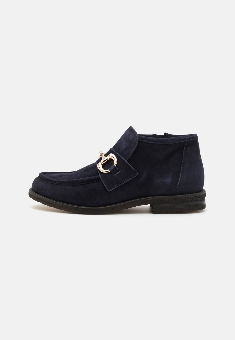Botas de ante azul marino hasta el tobillo con punta redonda, que cuentan con una hebilla dorada decorativa y una suela plana, mostrando una textura suave.