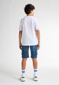 Camiseta blanca de manga corta, pantalones cortos de cargo vaqueros azules, zapatillas blancas con acentos verdes y calcetines blancos con rayas azules, visto desde atrás.