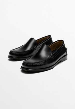 Paire de mocassins noirs en cuir à enfiler avec des détails cousus, bout arrondi et talon bas, présentés sur un fond blanc.