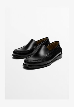 Paire de mocassins noirs en cuir à enfiler avec des détails cousus, bout arrondi et talon bas, présentés sur un fond blanc.