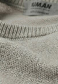 Jasnoszary dzianinowy materiał swetra z prążkowanym kołnierzem, ukazujący miękką fakturę i subtelne różnice kolorystyczne. W tle widoczna metka.