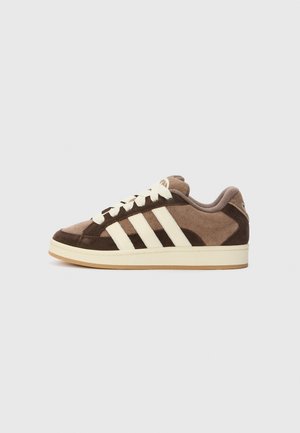 Bruine suède sneaker met witte veters en drie witte strepen aan de zijkant, in profiel getoond op een effen lichte achtergrond.