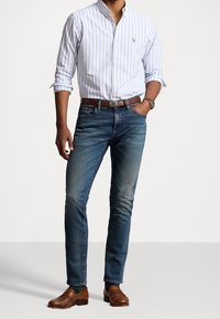 Herenoutfit met een lichtblauw en wit gestreept overhemd met knopen, donkere spijkerbroek, bruin leren instappers en een brown leren riem.