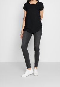 Camiseta negra de manga corta, tela suave, dobladillo curvado; combinada con jeans ajustados de color gris oscuro y zapatillas blancas con suelas texturizadas.