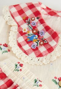 Red and white gingham collar with floral embroidery, lace trim. Cream fabric base with small floral patterns and gathered texture.