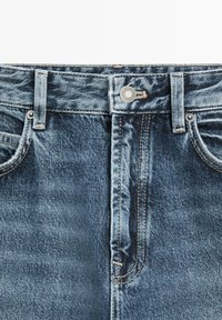 Close-up van de taille van een blauwe spijkerbroek met knoop, riemlussen, voorzakken en zichtbare stikseldetails.