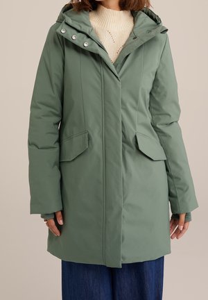 Vrouw draagt een winterjas met capuchon in saliegroen met voorvleugeltassen, over een crème gebreide trui en donkerblauwe broek.