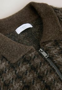 Brūns rāvējslēdzēja džemperis ar teksturētu, pūkainu materiālu. Iekļauj kontrastējošu melnu rakstu un platu apkakli. Redzams sudraba rāvējslēdzējs.