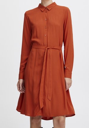 Robe-chemise à manches longues de couleur orange brûlé avec boutons sur le devant, col et ceinture à la taille, portée par une personne devant un fond uni.