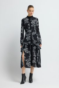 Schwarzes Kleid mit weißem Paisley-Muster, langen Ärmeln, hohem Kragen, Knopfleiste vorne, Rüschensaum und Seitenschlitzen; kombiniert mit schwarzen Stiefeletten.