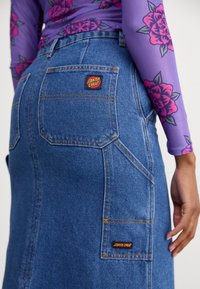 Jupe en denim avec plusieurs poches, de couleur bleu clair, et des coutures oranges contrastantes. Présente un patch logo "Santa Cruz" au dos.