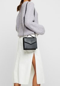 Mała czarna torba z eko-skóry z ćwiekami, trójkątną klapką i odpinanym paskiem, noszona do jasnej spódnicy z rozporkiem.