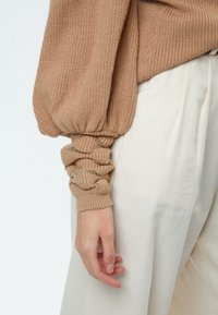 Beige gerippter Strickpullover mit Ballonärmeln und gerafften Bündchen, mit einem Akzent aus goldener Kettendetail am Handgelenk, kombiniert mit cremefarbenen Hosen.