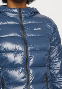 Modrá péřová bunda vyrobená z lesklého materiálu, s zipovým uzávěrem, kapucí a bočními kapsami. Logo "HUGO" na hrudi v bílé barvě.