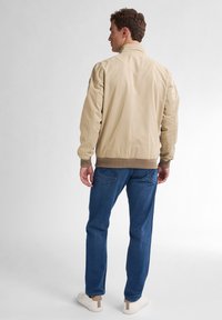 Jachetă bomber bej cu guler în picioare, manșete și talie ribbed. Design simplu, fără modele, asortată cu blugi albaștri și adidași albi.