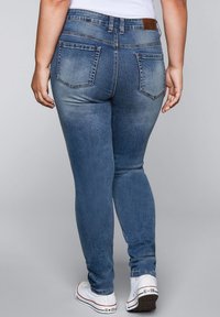 Úzké modré džíny z denimu se středním pasem, zadními kapsami, oprýskanými detaily a mírně vybledlým vzhledem, v kombinaci s bílými teniskami.