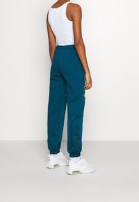 Pantalones de chándal en color verde azulado con puños elásticos, bolsillos laterales y acentos gráficos amarillos. Combinados con zapatillas deportivas blancas y una camiseta sin mangas blanca.