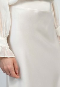 Jupe en satin couleur crème avec une texture lisse, présentant une taille haute et un ajustement près du corps, complétée par des manches en blouse blanche transparentes et froncées.