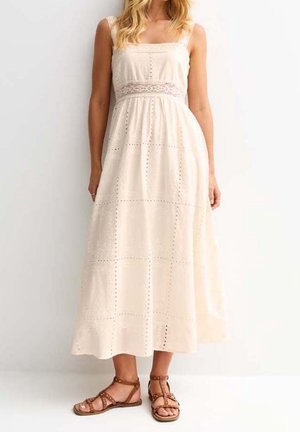 Femme portant une robe maxi crème sans manches avec des détails en œillets et des sandales plates marron cloutées, debout devant un fond blanc.
