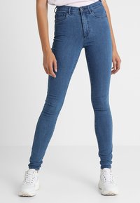 Jeans skinny a vita alta blu con una superficie in denim morbido. Presentano cinque tasche e una chiusura con bottone. Abbinati a sneakers bianche.