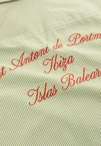 Tissu rayé vert et blanc avec une broderie cursive rouge indiquant "Sant Antoni de Portmany, Ibiza, Îles Baléares."