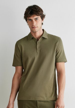 Mężczyzna ubrany w oliwkową koszulkę polo z krótkim rękawem stoi oparty o jasną, jednolitą ścianę, patrząc przed siebie z neutralnym wyrazem twarzy.