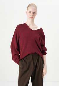Burgunderfarbener Oversize-Pullover mit tiefem V-Ausschnitt und gerippter Struktur, kombiniert mit hoch geschnittenen braunen Plisseehosen.