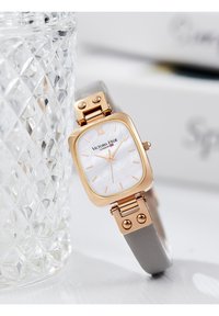 Montre rectangulaire avec un boîtier en acier inoxydable rose gold, un cadran en nacre, un bracelet en cuir gris et de solides accents métalliques.