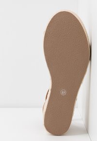 Semelle d'une sandale beige avec une texture antidérapante et la taille 37 indiquée, positionnée debout contre une surface d'angle blanche.