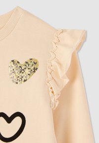 Felpa beige chiaro con un design di cuore dorato in paillettes e un'accennata arricciatura sulla spalla. Texture morbida, collo a coste lavorato a maglia.