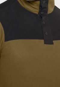 Sudadera de forro polar en verde oliva con una sección superior negra. Cuenta con un cuello alto y botones a presión. La textura es suave y lisa.