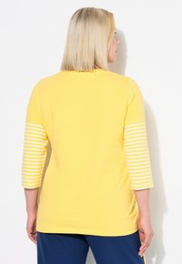 Gul topp med trekvarts randiga ärmar, mjukt bomullsmaterial, rundad nederkant och en enkel halsringning. Inga synliga grafiska motiv eller text.