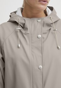 Beige regnjakke med hette, utstyrt med sølvfargede detaljer, snorjusteringer og trykknapper, laget av glatt, vannavstøtende materiale.