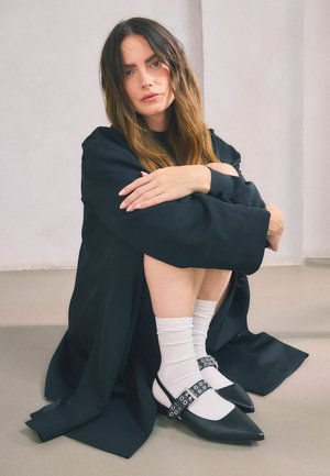 Scarpe nere e bianche a punta con una larga cinghia dotata di accessori in argento, indossate con calze bianche a coste. Modella seduta, indossa un cappotto nero.