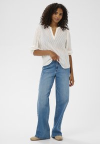 Femme aux cheveux bouclés portant un haut texturé blanc, un jean large bleu et des chaussures beiges, se tenait contre un fond uni.