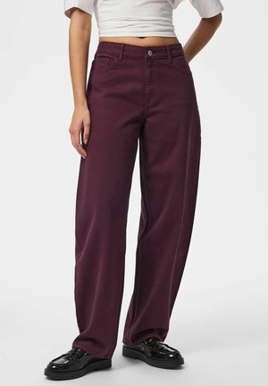 Person trägt dunkelviolette, hoch taillierte, weit geschnittene Hose kombiniert mit schwarzen Loafers und einem kurzen weißen Oberteil und steht vor einem schlichten Hintergrund.