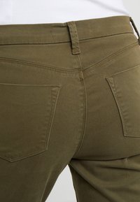 Olivgrön denimbyxor med en figurnära design, två bakfickor och synliga sömmar, som visar en slät, lätt texturerad yta.