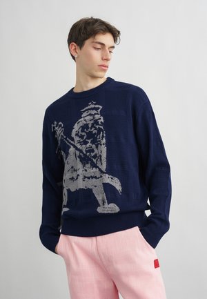 Nuori mies, jolla on yllään tummansininen neule harmaalla leijonakuvalla ja vaaleanpunaiset housut, seisoo kädet taskuissa yksinkertaisen taustan edessä.