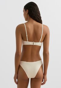 Dvodelne bikini v svetlo kremni barvi z teksturiranim vzorcem. Zgornji del ima nastavljive naramnice in zaponko na hrbtu; spodnji del je nizkega pasu.