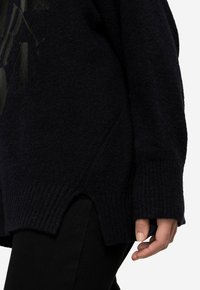 Dunkelblauer Strickpullover mit gerippten Bündchen, strukturierter Oberfläche und seitlichen Schlitzen am Saum. Eine Hand ruht auf dem Stoff.