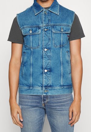 Mężczyzna w niebieskiej kamizelce dżinsowej na ciemnoszarej koszulce z krótkim rękawem i jasnoniebieskich jeansach, stojący na jednolitym tle.