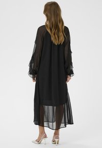 Kvinde med langt, bølget hår iført en gennemsigtig sort langærmet midikjole og hvide høje slingback-sko, står med ryggen synlig.