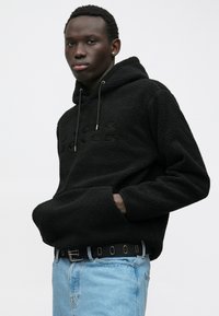 Νεαρός άνδρας που φοράει μια μαύρη υφασμάτινη κουκούλα με μπροστινή τσέπη και κουκούλα, ανοιχτούς μπλε τζιν και μια μαύρη ζώνη με μεταλλικά μάτια, σε λευκό φόντο.