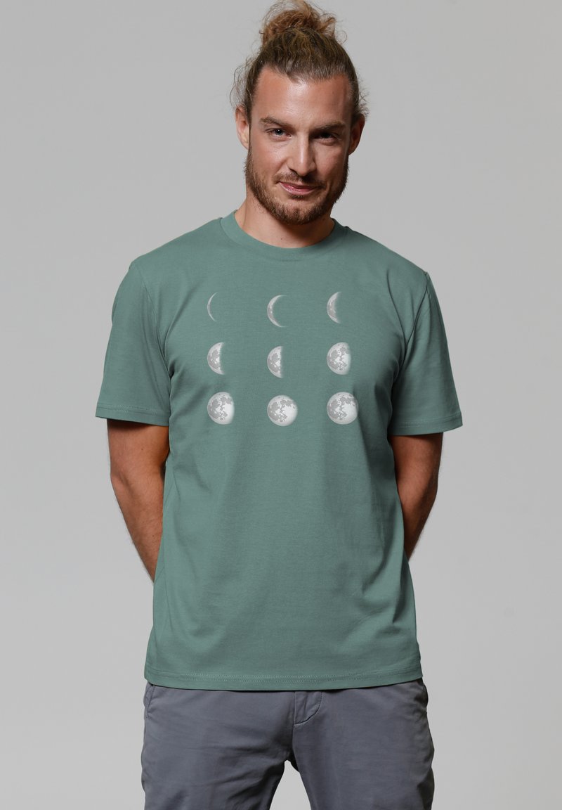 T-shirt en coton vert présentant un imprimé des phases lunaires en blanc, disposé en deux colonnes verticales, avec des manches courtes et un col rond.