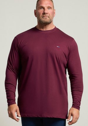 Uomo che indossa una maglia a maniche lunghe, color bordeaux scuro, con scollo tondo e jeans scuri, in piedi contro uno sfondo chiaro e uniforme, rivolto in avanti.