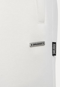 Pantalones jogger de tela blanca con una textura suave, que presenta una etiqueta metálica "SMILODOX" y una etiqueta negra en la costura lateral.