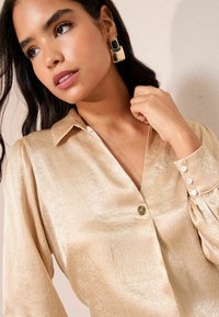 Helles, goldenfarbenes, strukturiertes Blusenshirt mit Kragen, Vorderknopfverschluss und Puffärmeln. Dekorative Knöpfe an den Ärmeln setzen Akzente.